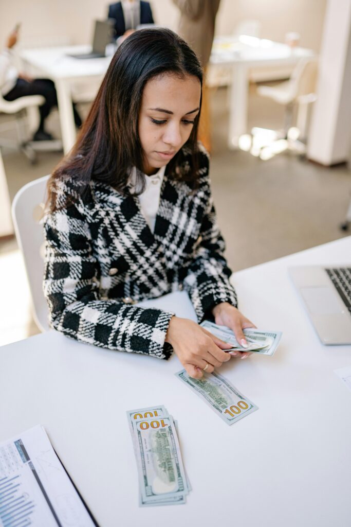 Female in plaid coat counting US dollar bills at office with laptop.